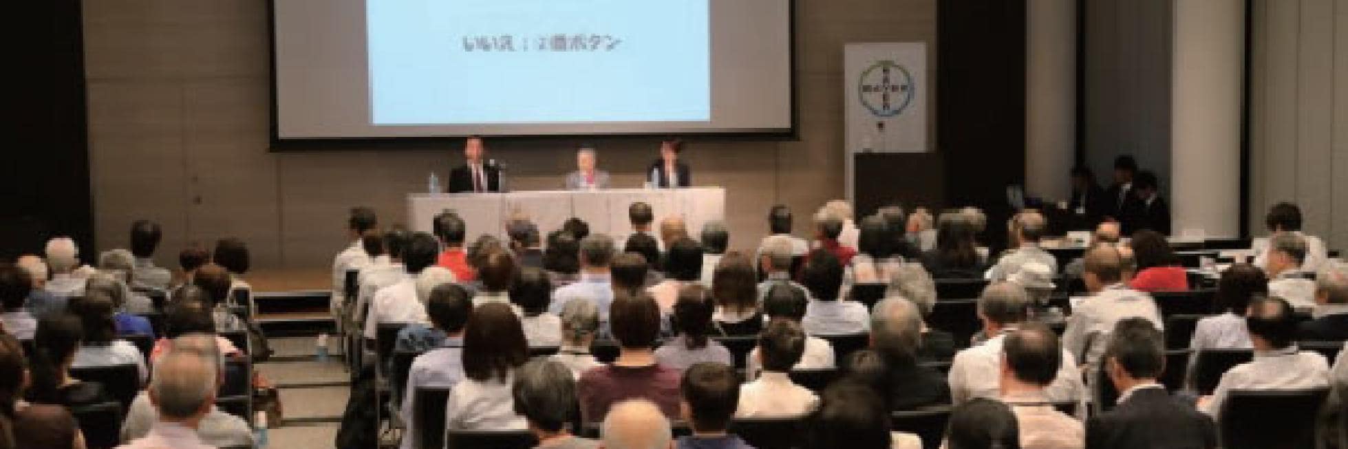 聴衆は会議室で、3 人の講演者と投影されたプレゼンテーション スクリーンがあるステージに向かって座っています。