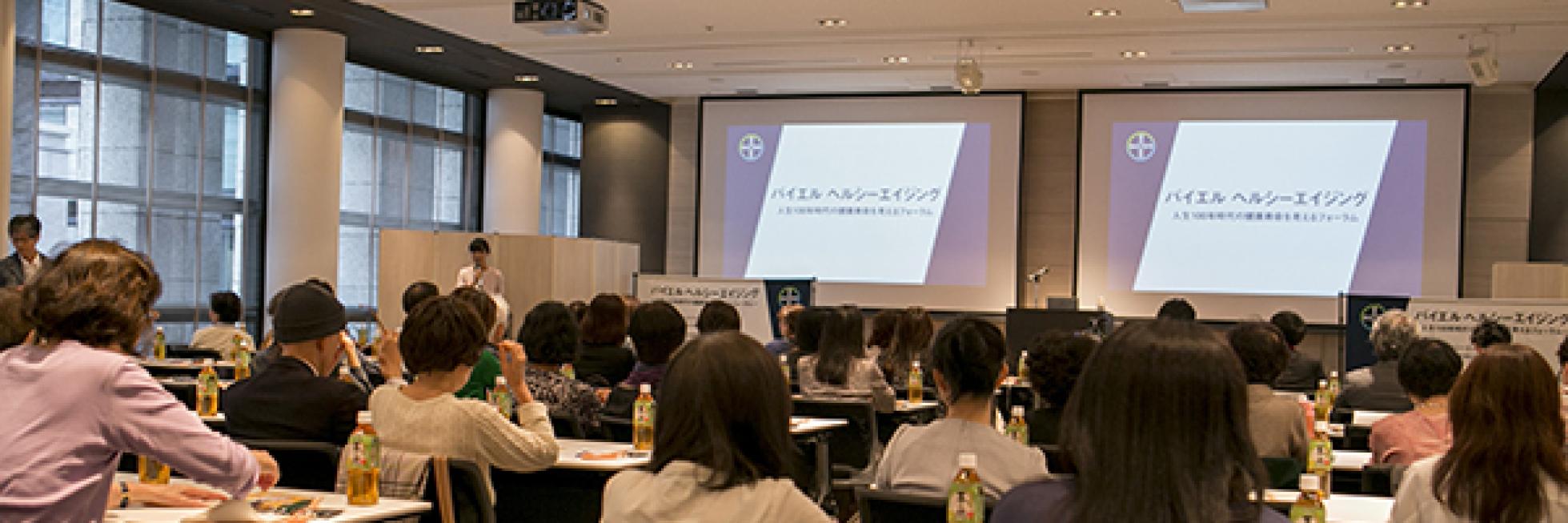 会議室には、プレゼンテーションのスライドが映し出された 2 つのプロジェクター スクリーンに向かって着席した出席者がいます。テーブルの上にはボトルと書類が置かれています。